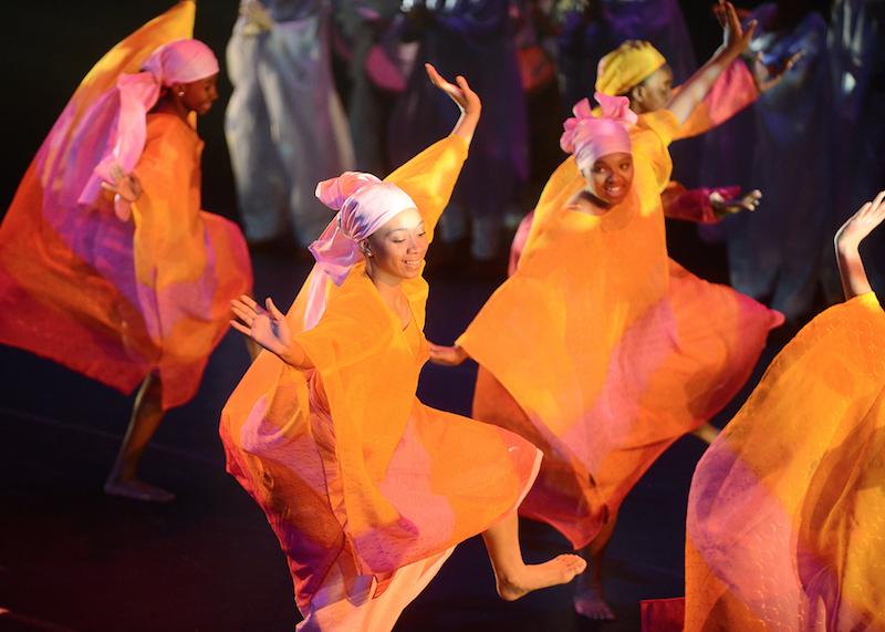 DanceAfrica Ensemble, Photography by  Dino Perrucci- smiling women dancing covered in colorful large silks of orange and magenta with equally beautiful head scarves 